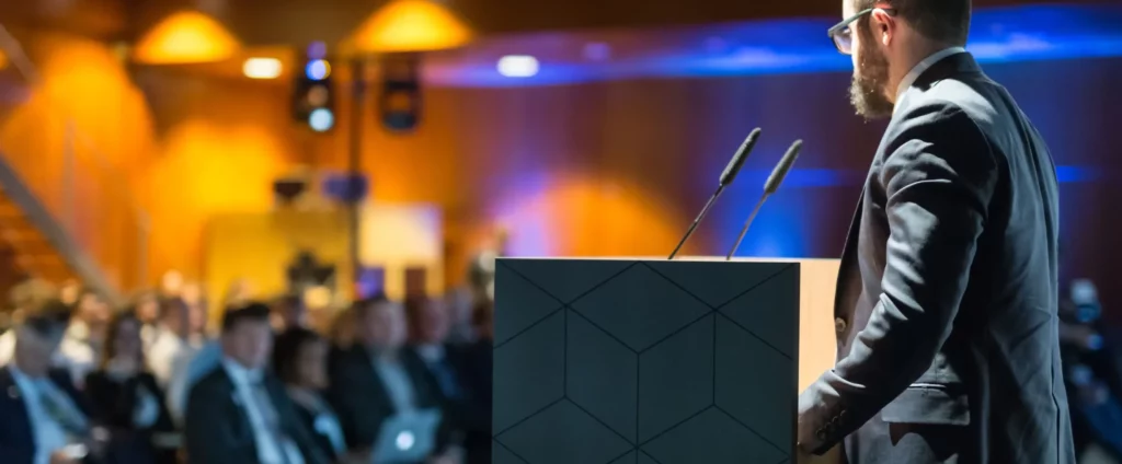 A speaker stands at a podium in front of an audience during a conference or presentation.