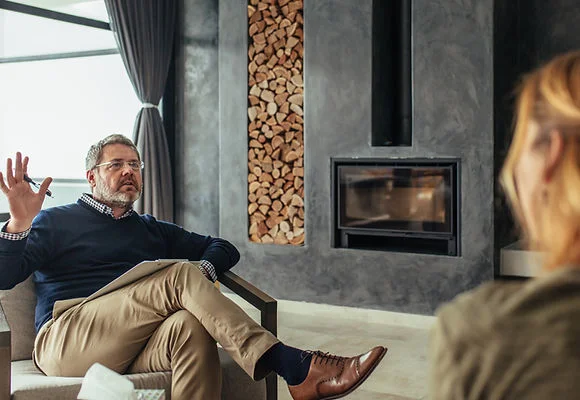 A man in a sweater sits in a modern room, gesturing while speaking to someone out of view.