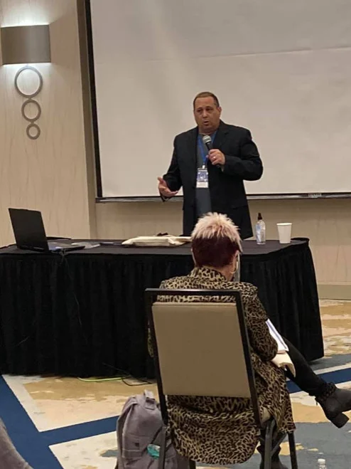 A speaker presents at a conference while an audience member listens from the front row.
